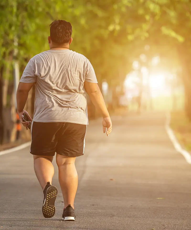 Overweight man walking outdoors on a sunny path, symbolizing the journey to improved fitness, health, and well-being through small, consistent actions. 2024. Mark Ludlow – The Life Changing Health Coach.