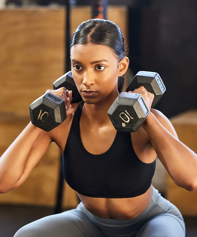 Focused woman performing a squat with dumbbells in a gym setting, representing empowerment through strength training and building sustainable fitness habits. 2024. Mark Ludlow – The Life Changing Health Coach.